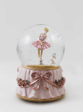 Load image into Gallery viewer, A decorative clear glass snow globe with a ballerina figurine in a pink tutu inside, set on a pale pink ruffled base with a large pink ribbon bow and small floral details, photographed against a light gray studio background.
