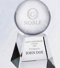 Load image into Gallery viewer, A clear glass or acrylic anniversary trophy with a round top engraved with a stylized G and the word NOBLE, mounted on a faceted pedestal. The front of the base is engraved with the message 'HAPPY ANNIVERSARY 5 YEARS 2026 Presented to JOHN DOE'.
